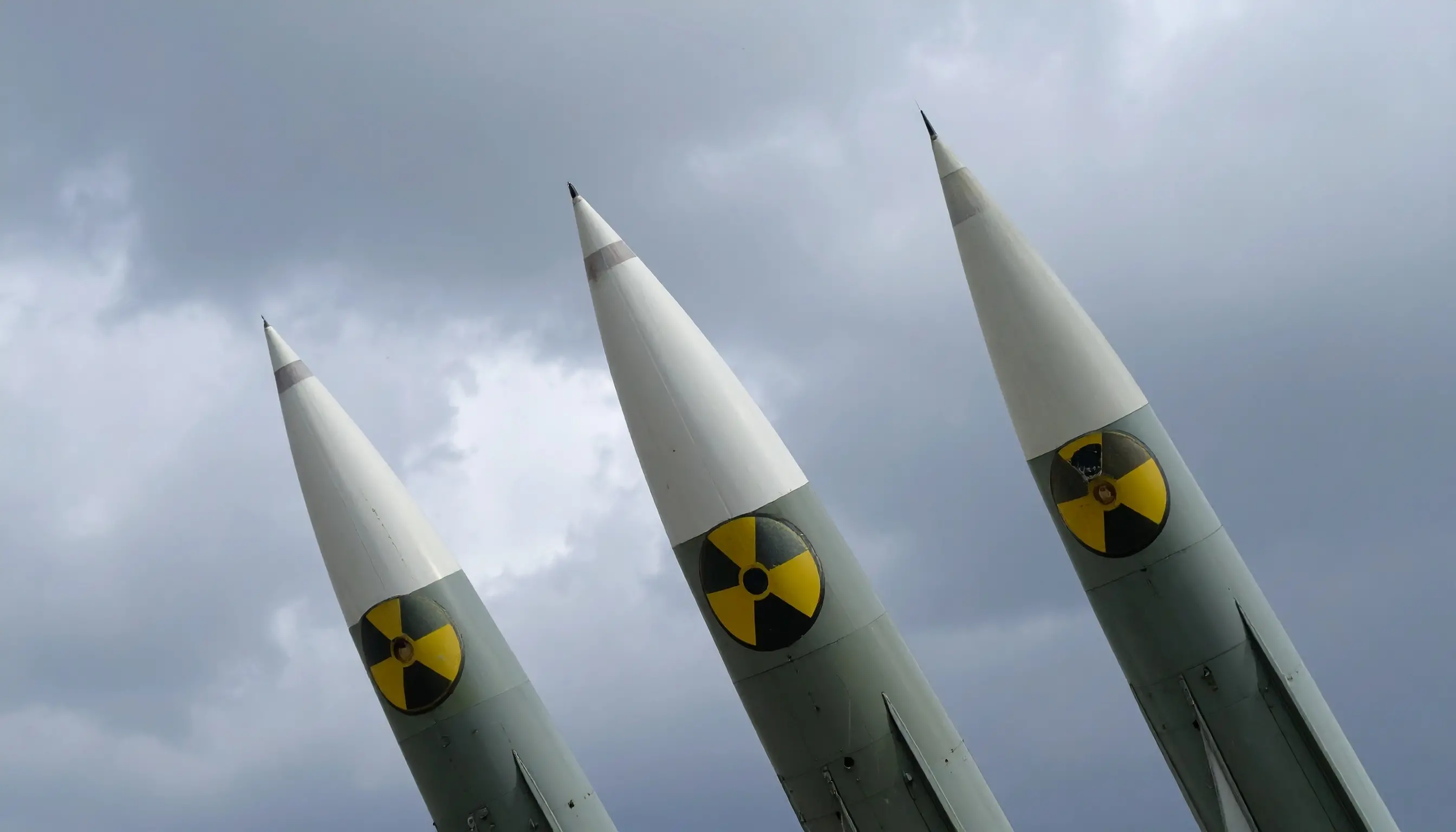 Three nuclear missiles pointed upward toward a cloudy sky. Each missile bears a radioactive hazard symbol. 